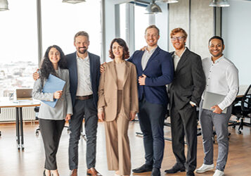 A group of six diverse business professionals stand together in a modern office setting. They are all smiling and appear to be in a good mood. The team is comprised of two women and four men